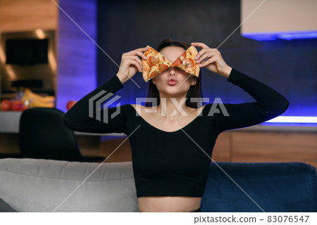 Excited happy young woman in kitchen plaing with pizza slices. Cover eyes with food. Carefree housekeeper very playful. Wear white dressing gown. Careless houseswife. 83076547