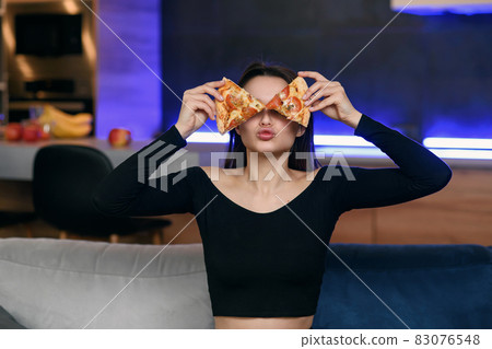 Excited happy young woman in kitchen plaing with pizza slices. Cover eyes with food. Carefree housekeeper very playful. Wear white dressing gown. Careless houseswife. 83076548