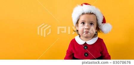 Banner Portraite Happy Cheerful Chubby Baby Girl in Santa Suit Looking Up Interested At Yellow Background. Child Play Christmas Scene Celebrating Birthday. Kid Have Fun Spend New Year Time Copy Space 83078676
