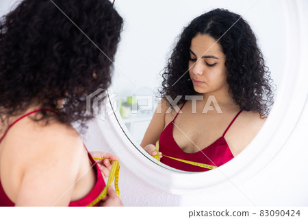 Girl measures her breasts with centimeter looking at reflection in the mirror 83090424