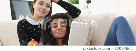 Caucasian and African American women read daily planner on sofa Caucasian and African American women read daily planner on sofa 83094663