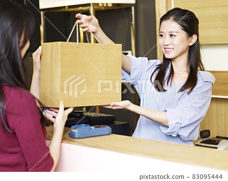 young asian salesclerk handing a paper bag of merchandise to a customer at the check-out counter young asian salesclerk handing a paper bag of merchandise to a customer at the check-out counter 83095444