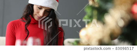 Sad woman in red santa claus hat sitting near two glasses of champagne and christmas tree Sad woman in red santa claus hat sitting near two glasses of champagne and christmas tree 83098517