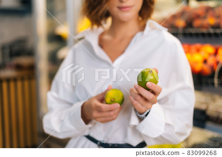 Close-up cropped shot of unrecognizable young woman holding in hands lime posing standing at fruit and vegetables section of grocery store. 83099268