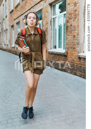 Teen schoolgirl on the way to the school. She is listening to music on headphones and holding a book. Education and back to school concept Teen schoolgirl on the way to the school. She is listening to music on headphones and holding a book. Education and back to school concept 83099779