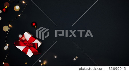 Holiday winter background. White gift box with red ribbon, New Year balls and sparkling lights garland in Christmas composition on black for greeting card. Flat lay, top view, copy space. Holiday winter background. White gift box with red ribbon, New Year balls and sparkling lights garland in Christmas composition on black for greeting card. Flat lay, top view, copy space. 83099943
