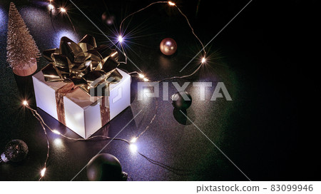 Surprise gift. White gift box with golden ribbon, New Year balls and sparkling lights garland in Christmas composition on black background for greeting card. Copy space. Surprise gift. White gift box with golden ribbon, New Year balls and sparkling lights garland in Christmas composition on black background for greeting card. Copy space. 83099946