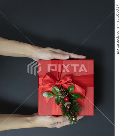 Hands hold a square red gift box with a decorative fir sprig on a black background. Top view. Christmas gift Hands hold a square red gift box with a decorative fir sprig on a black background. Top view. Christmas gift 83100317