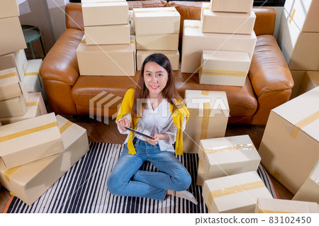 Young Asian woman hold tablet and sit among many boxes or carton and smile to camera 83102450