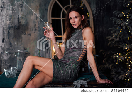 Young woman sits by Christmas tree with glass on eve of Christmas Young woman sits by Christmas tree with glass on eve of Christmas 83104483