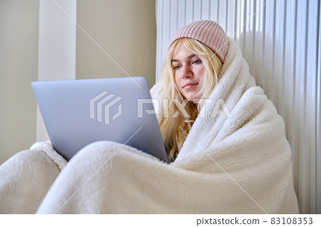 Teenager frozen in winter cold season, warming up with blanket hat, looking at laptop 83108353