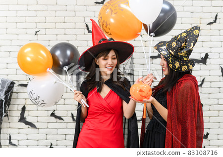 Two beautiful young asian woman in witch halloween costume holding pumpkin Jack O Lantern and balloon on white brick background decorated with black paper bat shape for halloween party Two beautiful young asian woman in witch halloween costume holding pumpkin Jack O Lantern and balloon on white brick background decorated with black paper bat shape for halloween party 83108716