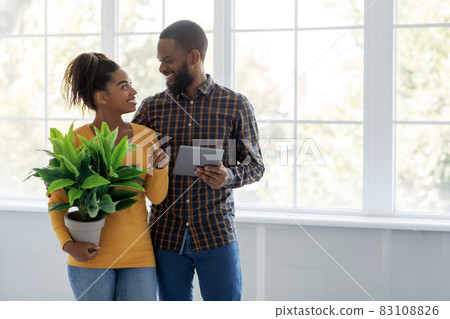 Cheerful millennial african american man and woman look at each other hold tablet and plant on window background Cheerful millennial african american man and woman look at each other hold tablet and plant on window background 83108826