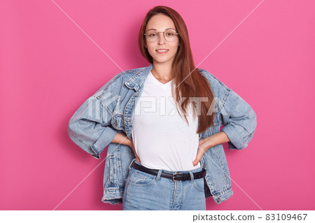 Close up portrait of young lady with denim clothing and spectacles, stands smiling isolated over pink studio background, keeps hands on hips, looking directly at camera. Fashion, people, jeans concept Close up portrait of young lady with denim clothing and spectacles, stands smiling isolated over pink studio background, keeps hands on hips, looking directly at camera. Fashion, people, jeans concept 83109467
