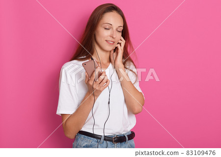 Portrait of beautiful girl in headphones listening to music with closed eyes, standing over pink studio background, wearing white casual t shirt, relaxes with favourite songs. Copy space for promotion 83109476
