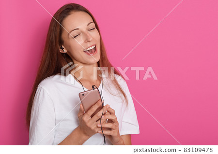 Image of happy carefree young woman dancing and listening to music from smartphone over pink background, lady wearing white shirt, holding cell phone. Copy space for advertisment or promotion text. 83109478