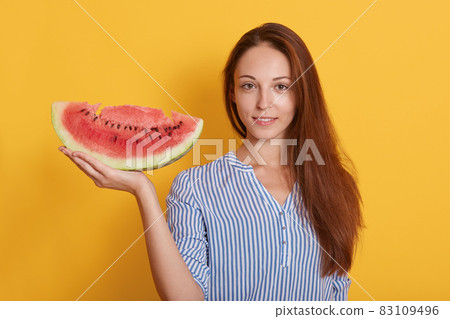 Image of young woman holding big slice of sweet watermelon while looking at camera, posing isolated over yellow background, adorable dark haired female having snack in hot summer day. People concept. Image of young woman holding big slice of sweet watermelon while looking at camera, posing isolated over yellow background, adorable dark haired female having snack in hot summer day. People concept. 83109496
