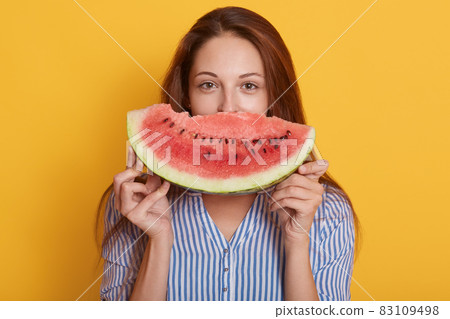 Horizontal shot of beautiful smiling young woman with slice of watermelon, female covering half of face with fruit, lady wearing blouse, having dark hair, posing isolated over yellow background. Horizontal shot of beautiful smiling young woman with slice of watermelon, female covering half of face with fruit, lady wearing blouse, having dark hair, posing isolated over yellow background. 83109498