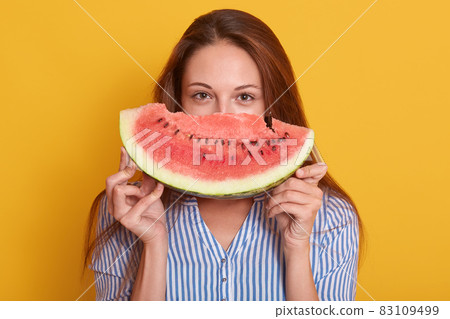 Image of young adorable lady dressed stylish striped shirt, holding piece of watermelon in hands, female hiding behind watermalon and looking directly at camera. People and summertime concept. 83109499