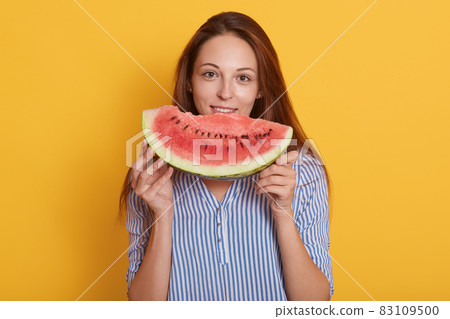 Close up portrait of beautiful brunette smiling young woman with slice of watermelon, charming lady wearing blouse posinhg isolated over yellow studio background, being in good mood, looks happy. Close up portrait of beautiful brunette smiling young woman with slice of watermelon, charming lady wearing blouse posinhg isolated over yellow studio background, being in good mood, looks happy. 83109500
