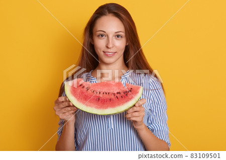 Horizontal shot of young woman with piece of watermelon in hands, female wearing striped shirt and having long brown hair, looking smiling at camera, model posing isolated over pink studio background. 83109501