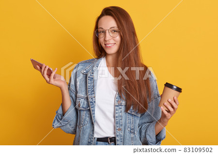 Studio shot of smiling young woman holding mobile phone and drinking take away coffee, surfing interner or chatting with friends, looking at camera, posing isolated over yellow studio background. Studio shot of smiling young woman holding mobile phone and drinking take away coffee, surfing interner or chatting with friends, looking at camera, posing isolated over yellow studio background. 83109502