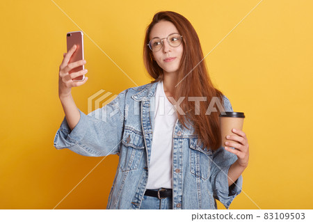 Portrait of pleasant tender lovely making selfie, holding smartphone and paper cup of coffee, wearing white t shirt and jeans jacket, having lunch, enjoying free time. Youth and rest concept. Portrait of pleasant tender lovely making selfie, holding smartphone and paper cup of coffee, wearing white t shirt and jeans jacket, having lunch, enjoying free time. Youth and rest concept. 83109503