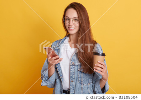 Picture of cheerful good looking young female standing isolated over yellow background in studio, holding paper cup with hot drink and smartphone, having delighted facial expression. Emotions concept. Picture of cheerful good looking young female standing isolated over yellow background in studio, holding paper cup with hot drink and smartphone, having delighted facial expression. Emotions concept. 83109504