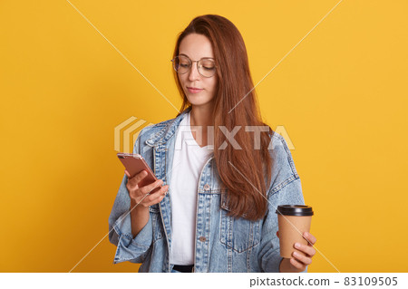 Studio shot of stupefied Caucasian woman reading news on internet website while drinking takeaway coffee, holding mobile phone, dresses white shirt and denim jacket, looks calm, poses indoor. Studio shot of stupefied Caucasian woman reading news on internet website while drinking takeaway coffee, holding mobile phone, dresses white shirt and denim jacket, looks calm, poses indoor. 83109505