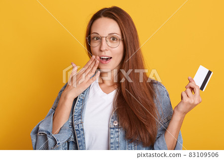 Shocked young Caucasian girl with long straight hair wearing denim jacket and white t shirt posing isolated over yellow background, holding credit bank card, keeps mouth opened, covering it with palm. Shocked young Caucasian girl with long straight hair wearing denim jacket and white t shirt posing isolated over yellow background, holding credit bank card, keeps mouth opened, covering it with palm. 83109506
