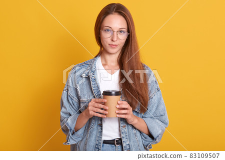 Studio portrait of cheerful lady looking directly at camera with pleasure, young woman holding hot coffee, wearing fashionable denim jacket, posing isolated over yellow background. Lifestyle concept. Studio portrait of cheerful lady looking directly at camera with pleasure, young woman holding hot coffee, wearing fashionable denim jacket, posing isolated over yellow background. Lifestyle concept. 83109507