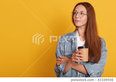 Close up portrait of pensive woman with dark hair, looking aside, contemplates about something, wearing white t shirt, denim jacket abd eyewear, holding disposable cup of coffee, over yellow wall. Close up portrait of pensive woman with dark hair, looking aside, contemplates about something, wearing white t shirt, denim jacket abd eyewear, holding disposable cup of coffee, over yellow wall. 83109508
