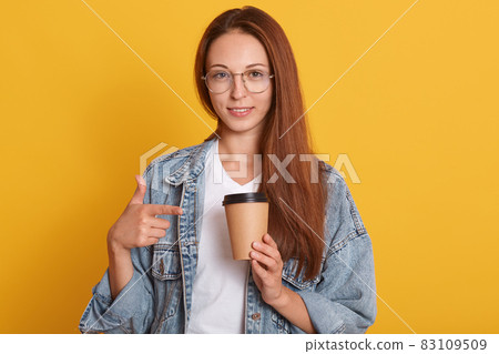 Image of young dark haired female pointing on paper coffee cup which holds in hand, woman dressed denim jacket and white t shirt, enjoying tasty hot takeaway baverage. People lifestyle concept. Image of young dark haired female pointing on paper coffee cup which holds in hand, woman dressed denim jacket and white t shirt, enjoying tasty hot takeaway baverage. People lifestyle concept. 83109509