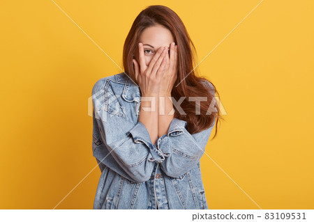 Portrait of cheerful young woman dresses denim casual jacket, covering her face with palms, posing isolated on bright yellow orange background in studio, expresses happyness. People lifestyle concept. Portrait of cheerful young woman dresses denim casual jacket, covering her face with palms, posing isolated on bright yellow orange background in studio, expresses happyness. People lifestyle concept. 83109531