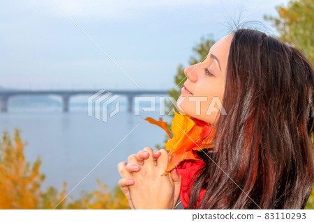 Pretty brunette girl holding yellow autumn leaves standing on a river shore. Pretty brunette girl holding yellow autumn leaves standing on a river shore. 83110293