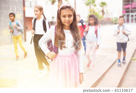 Positive schoolgirl standing near school, children on background Positive schoolgirl standing near school, children on background 83110617