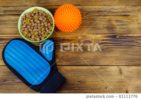 Canned dog food in bowl, ball toy and pet grooming glove on wooden background. Top view, copy space. Pet care concept 83111776