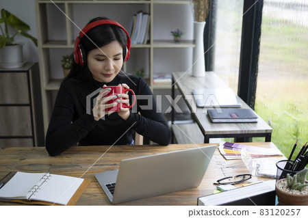 Young woman wearing headphone and drinking hot coffee in office. Young woman wearing headphone and drinking hot coffee in office. 83120257
