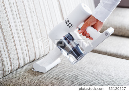 A person uses a white cordless vacuum cleaner to clean the sofa in the living room. 83120327