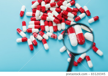 A handful of Red and white pill capsules are examined with a magnifying glass on a blue background 83120345