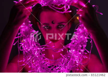 Woman with witch makeup and an earring in her nose. The girl holds a garland of flickering pink lights near her face. Light bulbs illuminate the witch's face in the dark. 83121462
