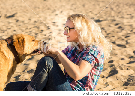 Portrait of young beautiful woman in sunglasses sitting on sand beach hugging golden retriever dog. Girl with dog by sea. Happiness and friendship. 83123353