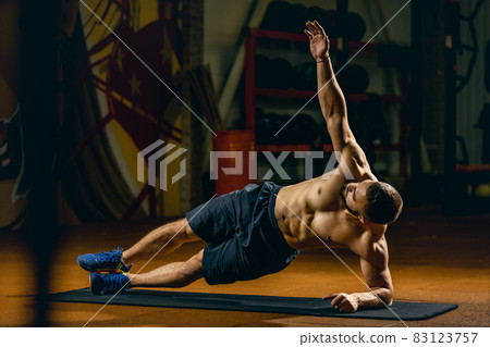 Full-length portrait of young sportive man standing in side plank position. Gym training Full-length portrait of young sportive man standing in side plank position. Gym training 83123757