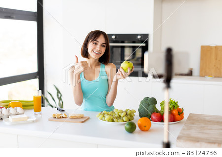 Happy woman showing thumb up and holding apple, recording new video for sport and food blog, kitchen interior Happy woman showing thumb up and holding apple, recording new video for sport and food blog, kitchen interior 83124736