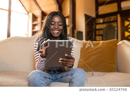 Happy young black lady using tablet, learning or working remotely, having online video call, sitting on sofa indoors Happy young black lady using tablet, learning or working remotely, having online video call, sitting on sofa indoors 83124748