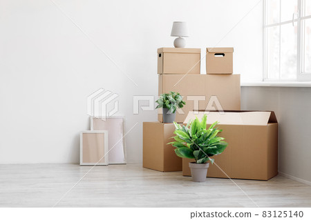 Stack of many different cardboard boxes with goods and green plants in pots on floor on white wall background 83125140