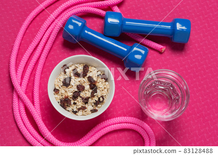 healthy lifestyle concept, on the fitness mat there is a bowl with muesli, two dumbbells, a jump rope and a glass of water 83128488