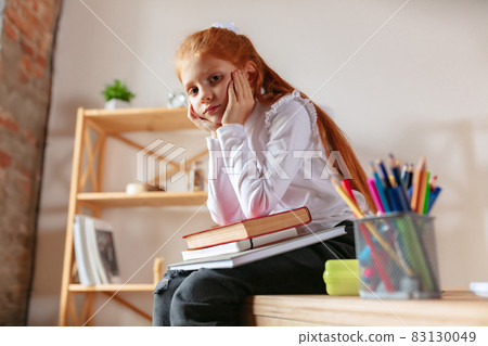 Portrait of little schoolgirl sitting on the table with lots of books. Online studying, homewrok tasks 83130049
