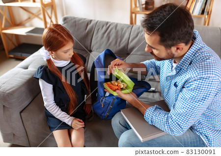 Top view image of responsible father helping his daughter to pack school backpack, putting lunch box Top view image of responsible father helping his daughter to pack school backpack, putting lunch box 83130091