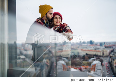 Young couple wrapped in blanket standing and hugging outdoors on balcony, shot through glass. Young couple wrapped in blanket standing and hugging outdoors on balcony, shot through glass. 83131652